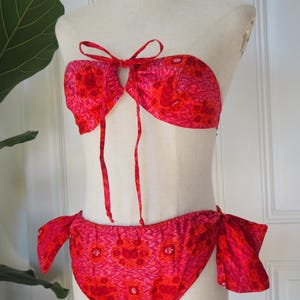 May include: A two-piece bikini set in shades of red and pink, displayed on a mannequin. The top is a bandeau style with a tie closure, and the bottoms have side ties. The fabric features a floral pattern.