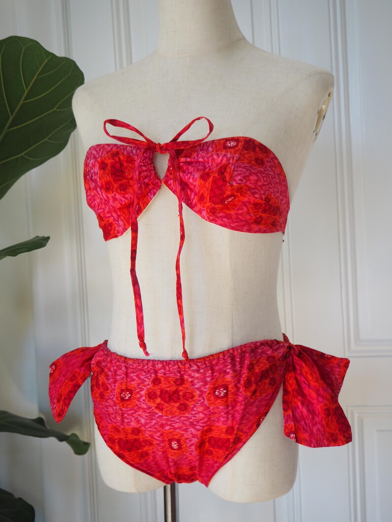May include: A two-piece bikini set in shades of red and pink, displayed on a mannequin. The top is a bandeau style with a tie closure, and the bottoms have side ties. The fabric features a floral pattern.