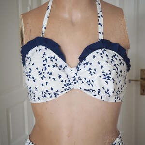 May include: A white halter-top bikini with a navy blue geometric pattern and navy blue ruffle trim. The bikini top is on a mannequin. The matching bottoms are visible at the bottom of the image. The bikini is made of a lightweight fabric.