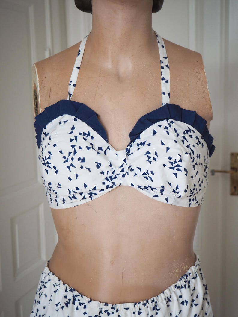 May include: A white halter-top bikini with a navy blue geometric pattern and navy blue ruffle trim. The bikini top is on a mannequin. The matching bottoms are visible at the bottom of the image. The bikini is made of a lightweight fabric.