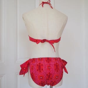 May include: A two-piece swimsuit in shades of red and pink, displayed on a mannequin. The halter top and the bottoms are adorned with a floral pattern. The bottoms have tie closures on each side. The swimsuit is made of a lightweight fabric.