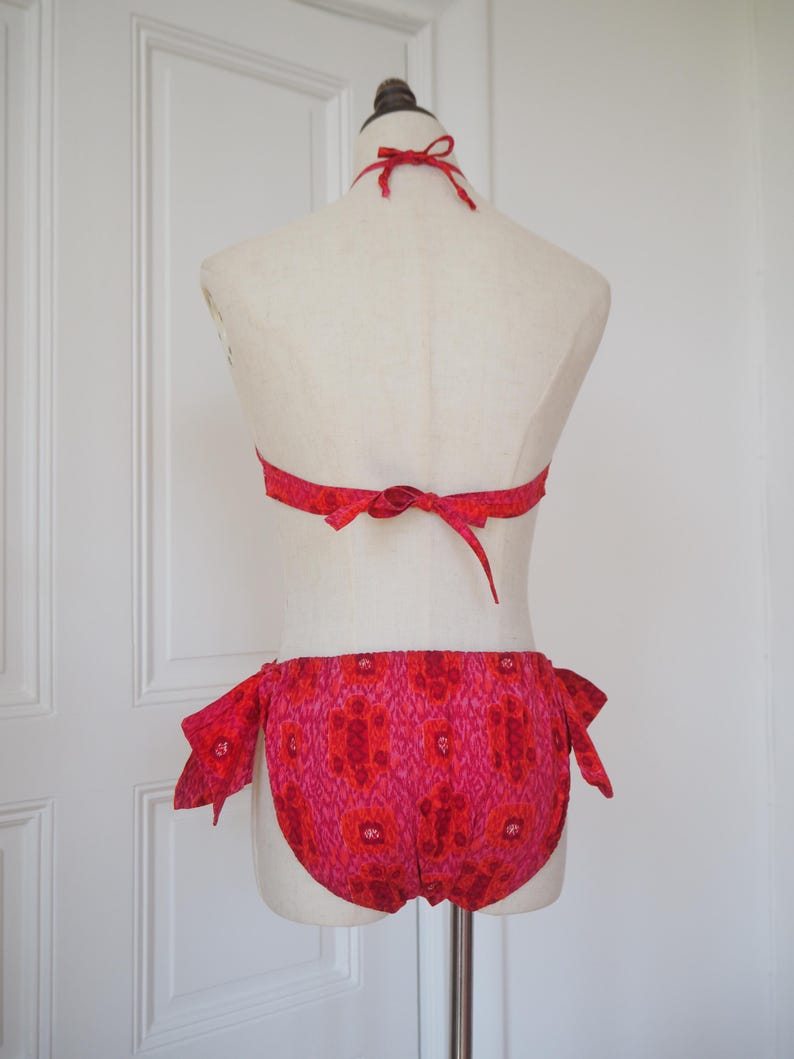 May include: A two-piece swimsuit in shades of red and pink, displayed on a mannequin. The halter top and the bottoms are adorned with a floral pattern. The bottoms have tie closures on each side. The swimsuit is made of a lightweight fabric.