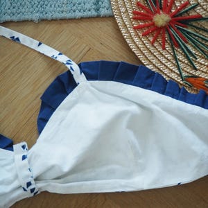 May include: A white and blue apron with a pleated navy blue top edge and white straps with blue triangle patterns. The apron is made of cotton and is laying on a wooden surface. A straw hat with red and green embellishments is in the background.