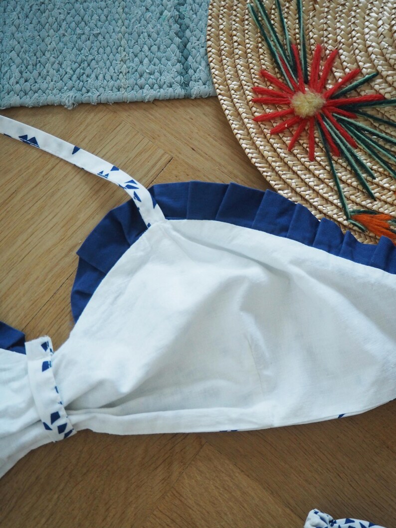 May include: A white and blue apron with a pleated navy blue top edge and white straps with blue triangle patterns. The apron is made of cotton and is laying on a wooden surface. A straw hat with red and green embellishments is in the background.