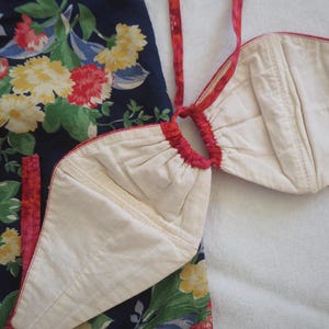 May include: A two-piece garment featuring a cream-colored top and a navy blue bottom with a floral pattern. The top has a halter-style design with red trim and straps. The floral pattern includes red, yellow, and pink flowers.