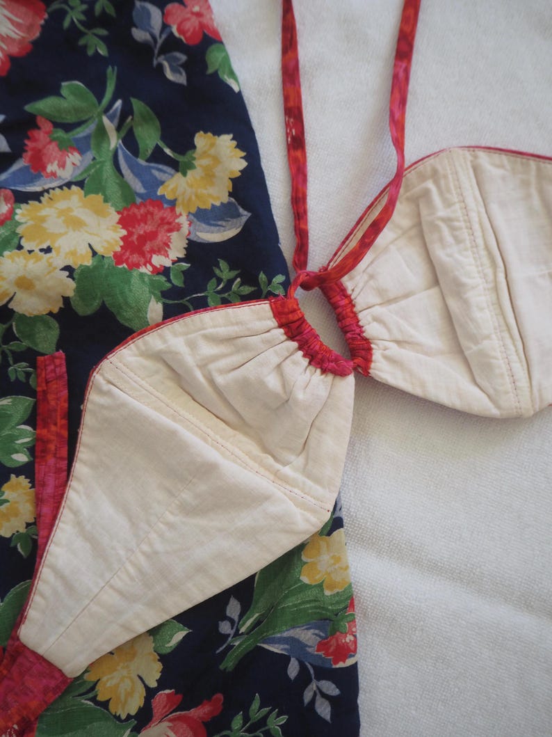 May include: A two-piece garment featuring a cream-colored top and a navy blue bottom with a floral pattern. The top has a halter-style design with red trim and straps. The floral pattern includes red, yellow, and pink flowers.