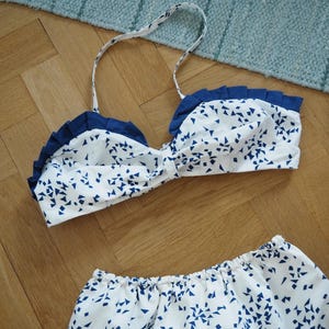 May include: A white and blue patterned bikini set. The top has a ruffled edge and thin straps. The bottoms are a matching pair of shorts. The pattern consists of small blue triangles scattered across a white background. The set is displayed on a wooden surface.