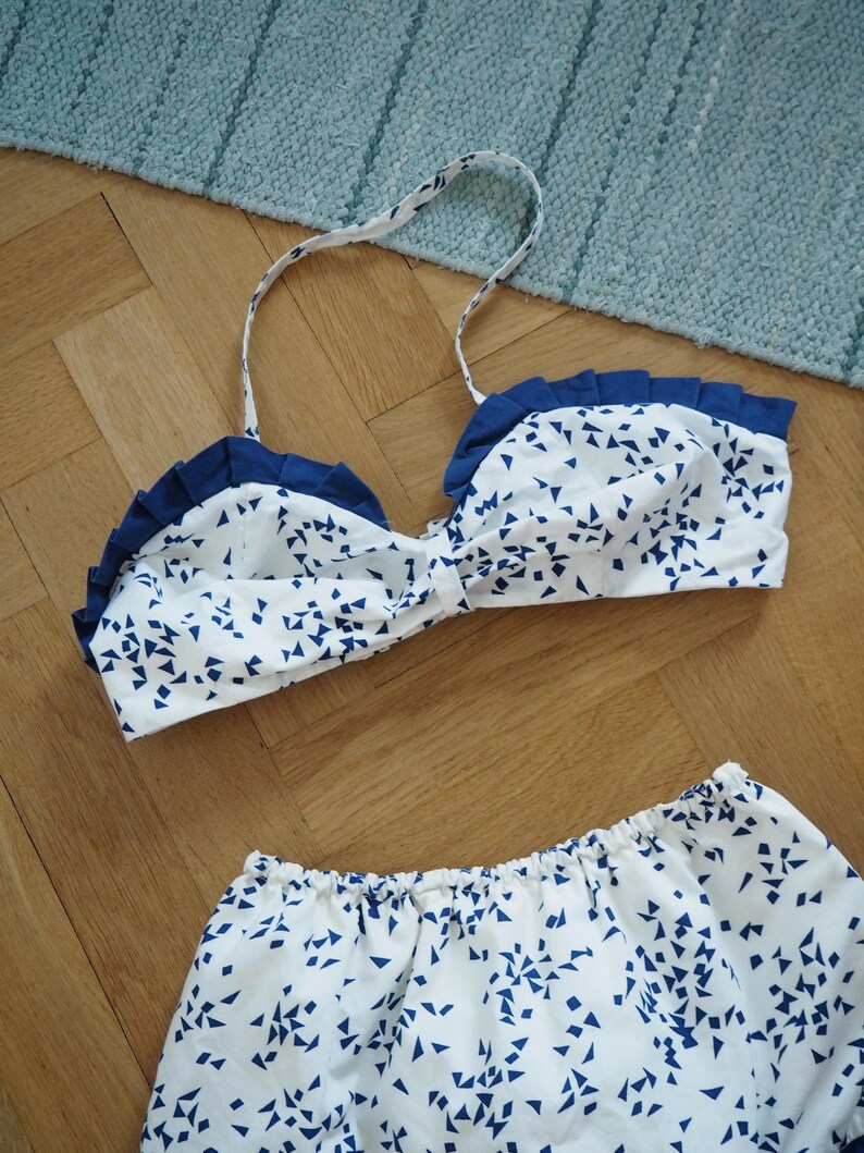May include: A white and blue patterned bikini set. The top has a ruffled edge and thin straps. The bottoms are a matching pair of shorts. The pattern consists of small blue triangles scattered across a white background. The set is displayed on a wooden surface.
