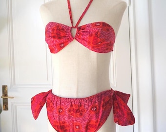 May include: A two-piece swimsuit in shades of red and pink, displayed on a mannequin. The top is a halter style with a keyhole detail, and the bottoms are a low-rise design with side ties. The fabric features a floral pattern.