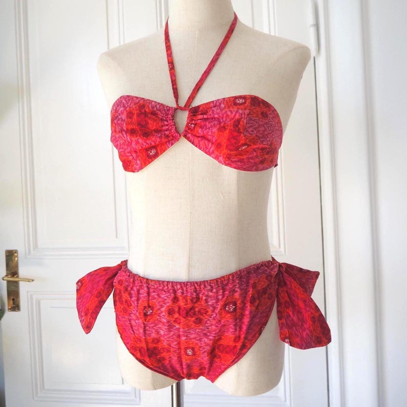 May include: A two-piece swimsuit in shades of red and pink, displayed on a mannequin. The top is a halter style with a keyhole detail, and the bottoms are a low-rise design with side ties. The fabric features a floral pattern.
