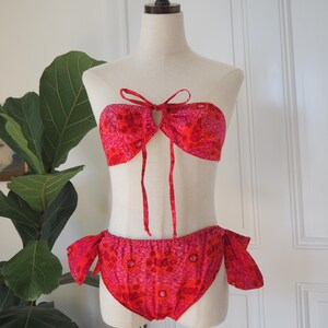 May include: A two-piece bikini set in shades of red and pink, displayed on a mannequin. The top is a bandeau style with a keyhole and tie closure. The bottoms are a classic cut with side ties. The fabric features a floral pattern.