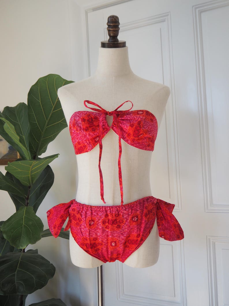 May include: A two-piece bikini set in shades of red and pink, displayed on a mannequin. The top is a bandeau style with a keyhole and tie closure. The bottoms are a classic cut with side ties. The fabric features a floral pattern.