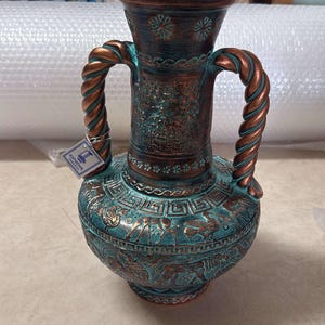 Puede incluir: Un jarrón ánfora de cerámica con acabado en color verde azulado y cobre, con dos asas trenzadas. El cuerpo está decorado con patrones griegos en relieve y motivos animales. Una pequeña etiqueta blanca muestra el texto KAPIATAK CERAMICS.