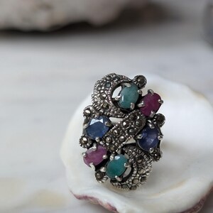 May include: A silver ring featuring a cluster of oval-cut gemstones including blue sapphires, red rubies, and green emeralds, accented with small, dark, faceted marcasite stones set in a swirling, textured metal frame with a polished finish.