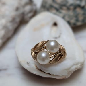 May include: A gold ring featuring two round white pearls set within a textured metal band designed with leaf-like patterns. The metal has a brushed finish, and the pearls are positioned side-by-side in the center of the decorative gold setting.