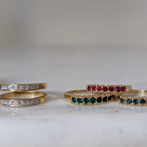 May include: A collection of gold-toned rings featuring various gemstone accents. One ring has a double band with clear stones, while three other rings are single bands set with rows of red, green, and blue stones respectively.