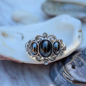 May include: A silver-toned ring featuring three oval-shaped, polished black stones set in a decorative, filigree-style metal frame. The central stone is larger than the two flanking stones, and the metalwork includes small, circular accents.