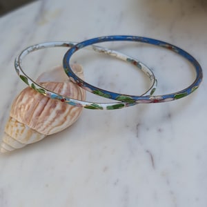 May include: Two thin metal bangles featuring colorful enamel floral and leaf patterns. One bangle has a white base with green, blue, and red accents, while the other features a vibrant blue base with similar floral motifs and metallic detailing.