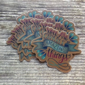 May include: A stack of brown paper stickers with blue and teal accents. The stickers feature the text "You Are Capable of Amazing Things" in a handwritten font.