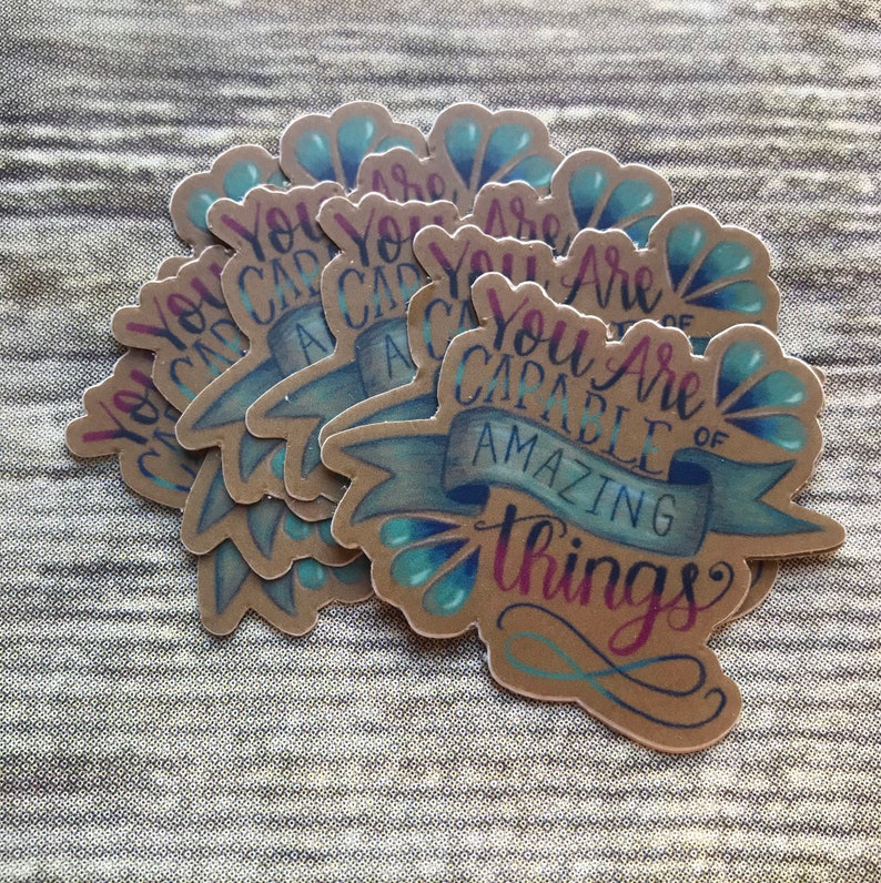 May include: A stack of brown paper stickers with blue and teal accents. The stickers feature the text "You Are Capable of Amazing Things" in a handwritten font.