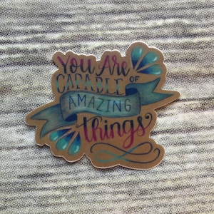 May include: A brown paper sticker with a blue ribbon and blue teardrop shapes. The sticker says "You are capable of amazing things" in a handwritten font.