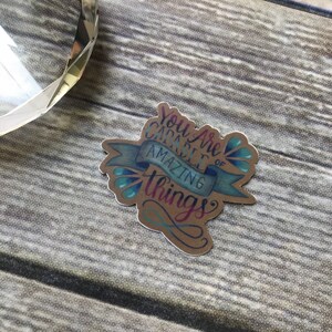 May include: A brown paper sticker with a blue ribbon and blue and green water droplets. The sticker says "You are capable of amazing things".