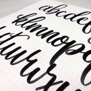 May include: A close-up of a handwritten alphabet in black ink on white paper with a dotted pattern. The letters are written in a cursive style.
