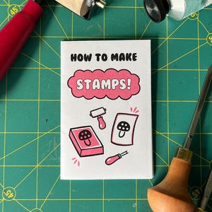 May include: A white booklet with the text "HOW TO MAKE STAMPS!" and illustrations of stamp-making tools and a mushroom design. A red roller, carving tools, and a tube of ink are also visible on a green cutting mat.