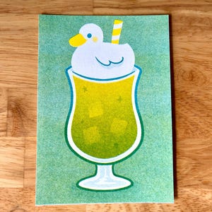 May include: Greeting card featuring a cartoon illustration of a green cocktail with ice cubes, a white duck-shaped foam, and a yellow striped straw. The background is a light green color.