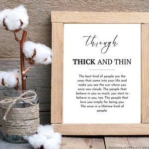 May include: A framed print with the words "Through Thick and Thin" in black script. A glass jar wrapped in twine holds cotton stems. The background is a rustic wooden surface.