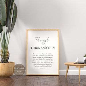 May include: A framed print with the words "Through Thick and Thin" in black script and text. The print is displayed on a wooden floor next to a potted plant and a small wooden table with a decorative bird and plant.