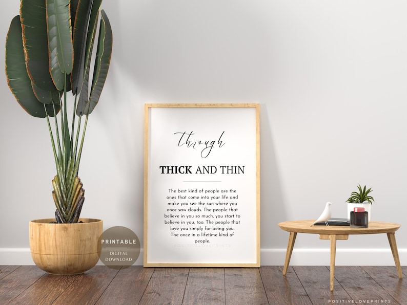 May include: A framed print with the words "Through Thick and Thin" in black script and text. The print is displayed on a wooden floor next to a potted plant and a small wooden table with a decorative bird and plant.