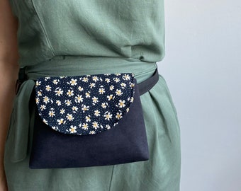 May include: A black fanny pack with a white daisy print flap. The fanny pack is worn around the waist of a person wearing a green dress.
