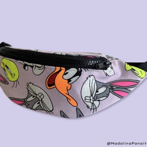May include: A purple fanny pack with a black zipper and a print of cartoon characters, including Bugs Bunny, Daffy Duck, Tweety Bird, and Sylvester the Cat.