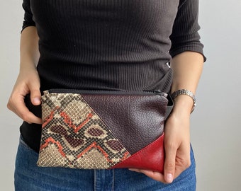 May include: A brown, red, and beige snakeskin-patterned fanny pack with a zipper closure. The fanny pack is worn around the waist of a person.