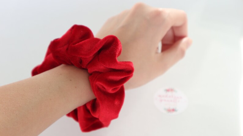 May include: A red velvet scrunchie, a type of hair tie, is shown on a person's wrist.