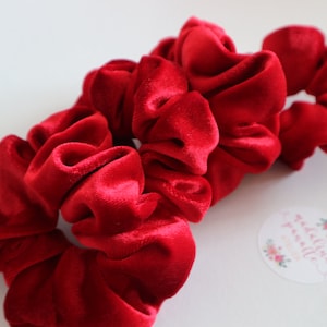 May include: Three red velvet scrunchies, a popular hair accessory, are stacked on top of each other. The scrunchies are made of soft, plush velvet and have a gathered, ruffled appearance.
