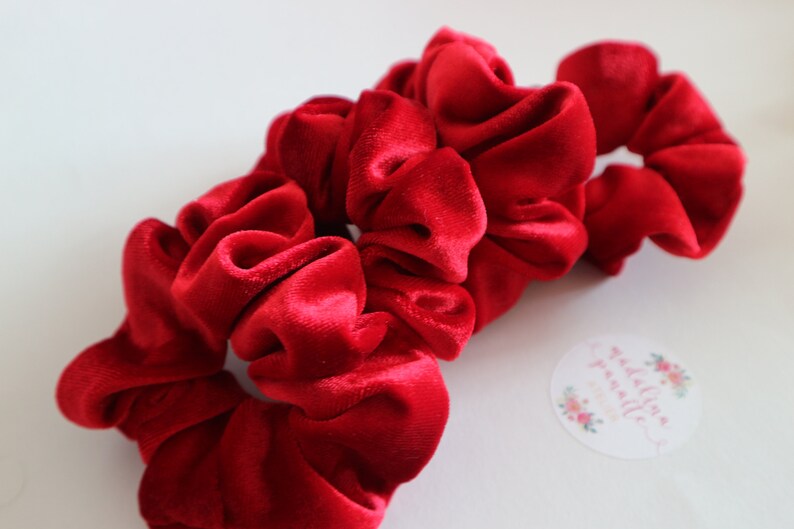 May include: Three red velvet scrunchies, a popular hair accessory, are stacked on top of each other. The scrunchies are made of soft, plush velvet and have a gathered, ruffled appearance.
