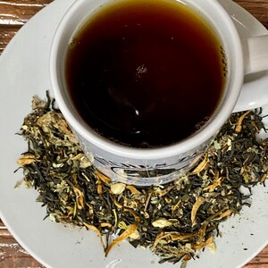 May include: A white ceramic teacup filled with dark brown tea, sitting on a white saucer with a scattering of loose leaf tea. The tea leaves are a mix of green, brown, and yellow.