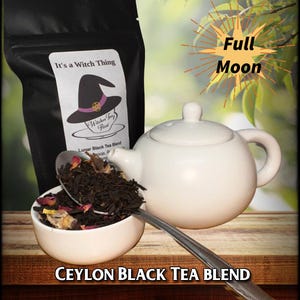 May include: A white ceramic teapot with a black tea blend in a bowl. The tea blend is labeled "Lunar Black Tea Blend" and "It's a Witch Thing". The tea bag is labeled "Witches Fey Flint". The text "Full Moon" is in the background.
