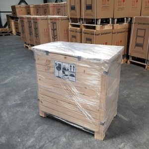 May include: A wooden crate with a clear plastic wrap, secured with black straps. The crate has a label with a symbol indicating fragile contents and arrows indicating the direction to lift the crate.