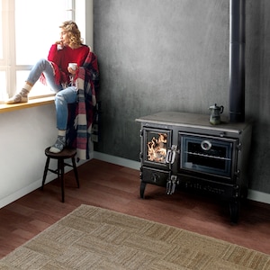 May include: A black cast iron wood-burning stove with a glass door and an oven. The stove has a decorative design on the front and a pipe extending from the top. The stove is sitting on a wooden floor with a rug in front of it.
