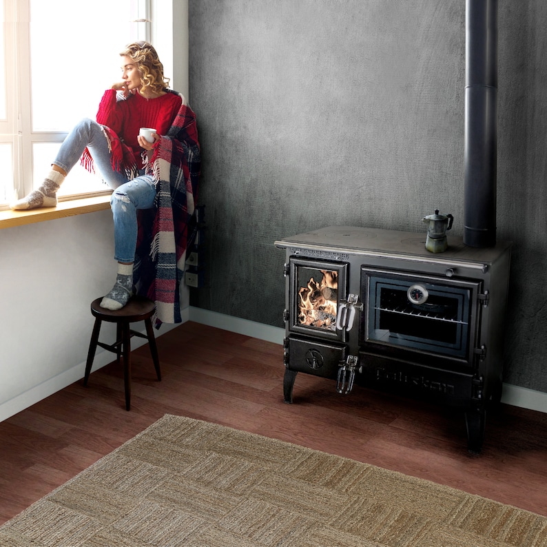 May include: A black cast iron wood-burning stove with a glass door and an oven. The stove has a decorative design on the front and a pipe extending from the top. The stove is sitting on a wooden floor with a rug in front of it.