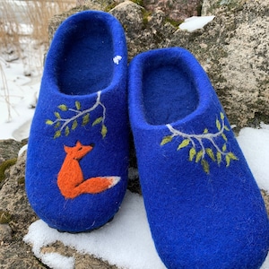 May include: A pair of blue felt slippers with a fox and leaf design. The slippers are a vibrant blue color, with an orange fox and green leaves embroidered on the front. The slippers are sitting on a stone surface with snow.