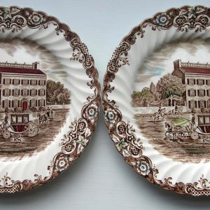 May include: Two round ceramic plates featuring a white center with a brown and multi-colored transferware scene of a large house, a horse-drawn carriage, and figures. The scalloped edges have a brown floral and lattice pattern border.