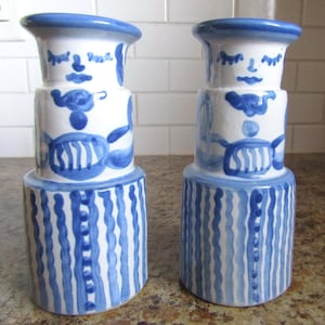 Puede incluir: Dos candelabros de cerámica blanca con diseños pintados a mano en azul. Cada pieza presenta una base cilíndrica con rayas onduladas verticales, una sección central con formas abstractas y una parte superior que representa un rostro estilizado con ojos cerrados.
