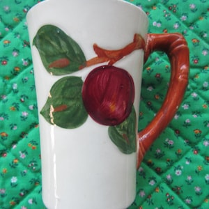 May include: White ceramic mug with a red apple and green leaf design. The handle is a reddish-brown color. The mug is a tall, cylindrical shape.