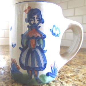 May include: A white ceramic mug featuring a hand-painted blue and orange illustration of a figure wearing a dress. The design includes small blue heart shapes and green ground details. The mug has a thick handle and a blue rim around the top edge.