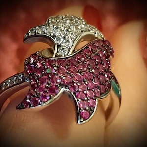 Peut inclure: Bague en argent ornée d'un motif d'aigle stylisé. La tête de l'aigle est incrustée de pierres précieuses transparentes, tandis que le corps est recouvert de petites pierres rose foncé. La bande de la bague est également ornée de pierres précieuses transparentes.