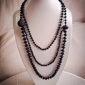 May include: A three-strand black beaded necklace displayed on a white jewelry bust. The necklace features small, round beads and larger, faceted beads, with a decorative cluster on one strand. The beads have a glossy finish.